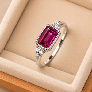 May include: A silver-toned ring featuring a rectangular, emerald-cut vibrant pink gemstone as the centerpiece. The stone is framed by a beaded metal border and accented with three small, clear round stones on each side of the band.
