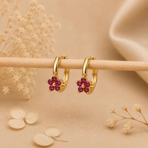 Puede incluir: Un par de pendientes de aro dorados, cada uno con un pequeño dije en forma de flor engastado con cinco piedras circulares de color rojo vibrante. Los pendientes cuelgan de una fina varilla de madera, mostrando el metal dorado y los pétalos rojos.