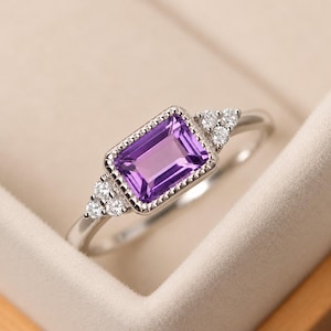 Puede incluir: Un anillo de tono plateado con una amatista morada rectangular de corte esmeralda en el centro, enmarcada por un borde de cuentas. Tres pequeñas piedras blancas redondas y transparentes están engastadas a cada lado de la amatista central.