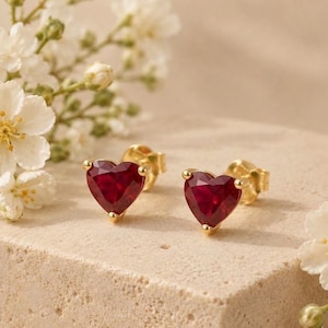 May include: A pair of stud earrings featuring heart-shaped red gemstones set in gold metal. Each earring has a faceted surface that reflects light, secured with a gold post backing. The design is simple, focusing on the vibrant red color of the stones.