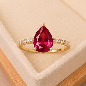 May include: A gold ring featuring a large, pear-shaped, deep red gemstone as the centrepiece. The band is crafted from polished yellow gold and is accented with a row of small, clear, round-cut stones embedded along both sides of the shank.