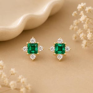 May include: A pair of gold stud earrings featuring a central square-cut green gemstone surrounded by four small round white stones arranged in a cross pattern. The metal setting is yellow gold, and the design highlights a symmetrical floral shape.