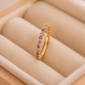 Op de afbeelding: Een gouden ring met een enkele rij kleine, ronde, paarse edelstenen die langs de bovenste helft van de band zijn gezet. De ring heeft een gepolijste metalen afwerking en staat rechtop om de gelijkmatige plaatsing van de stenen te tonen.