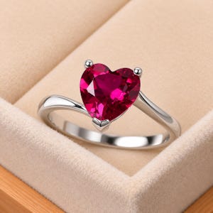 May include: A silver-toned ring featuring a prominent heart-shaped, deep red gemstone set in a three-prong setting. The band is smooth and polished, curving slightly to meet the base of the heart-shaped stone, creating a clean and simple design.