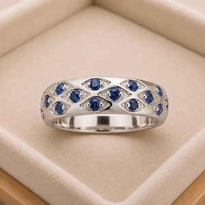Puede incluir: Un anillo de banda plateada con un patrón repetitivo de engastes metálicos en forma de marquesa, cada uno con una piedra preciosa redonda de color azul intenso. El diseño crea un motivo continuo e interconectado similar a ojos alrededor.