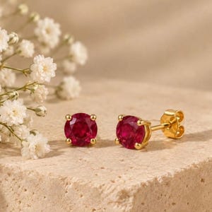 Puede incluir: Un par de pendientes de botón con piedras preciosas redondas, facetadas y de color rojo intenso, engastadas en oro amarillo pulido. Cada pendiente tiene un cierre de presión, mostrando un tono carmesí vibrante y un engaste clásico de garras.