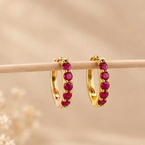 May include: A pair of gold hoop earrings featuring a row of small, round, vibrant red rubies set along the front-facing curve of each hoop. The earrings are suspended on a thin, horizontal wooden rod, showcasing the polished gold metal finish.
