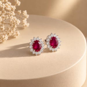May include: A pair of stud earrings featuring oval-cut deep red rubies surrounded by a halo of small, clear, round-cut white stones set in a bright silver-toned metal. The earrings are displayed together, showcasing the vibrant red centre stones.