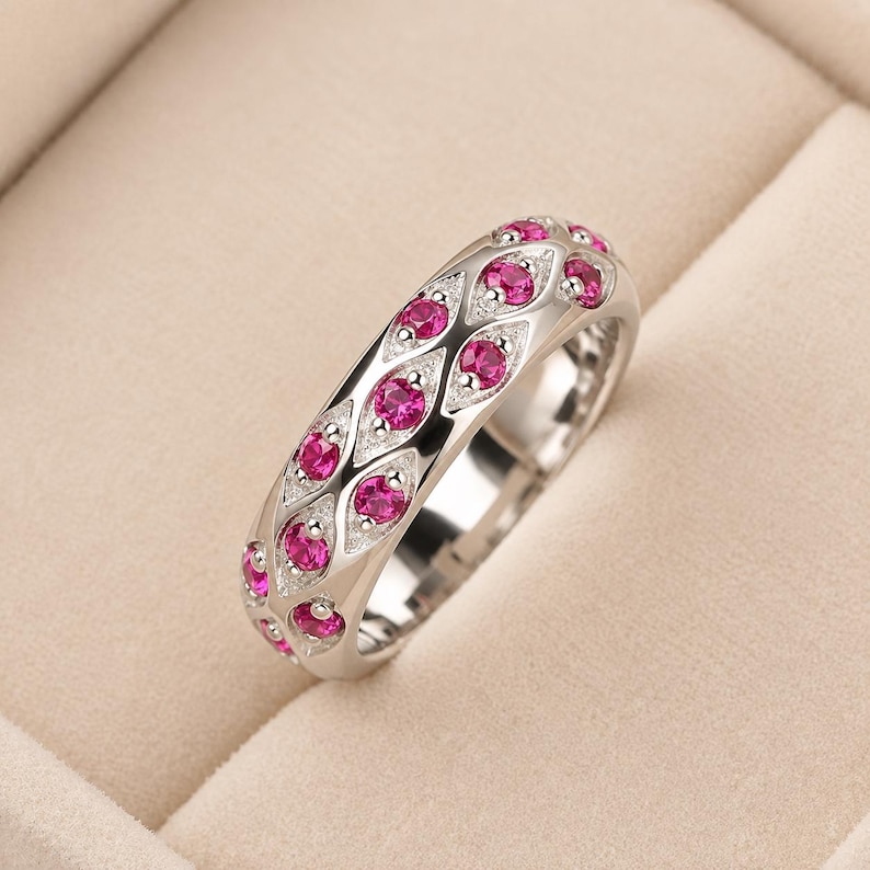 Puede incluir: Un anillo de banda en tono plateado con dos filas de engastes en forma de marquesa, cada uno con una peque&ntilde;a piedra preciosa redonda de color rosa vibrante. La superficie met&aacute;lica que rodea las piedras tiene un acabado granulado y texturizado.