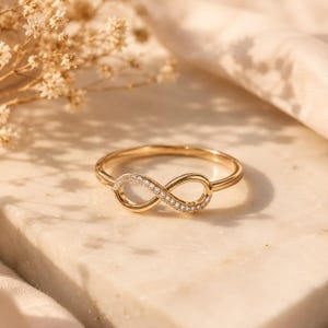 May include: A gold ring featuring an infinity symbol centrepiece. The infinity loop is adorned with a series of small, clear, round-cut stones embedded along one side of the metal band, creating a delicate and continuous design element.