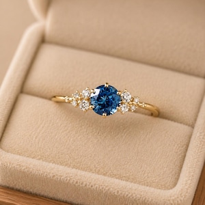 Puede incluir: Un anillo de oro con una piedra central redonda de color azul intenso, flanqueada por un grupo de tres piedras redondas más pequeñas y transparentes a cada lado. La banda es fina y de oro pulido, con un diseño delicado que resalta el contraste entre la vibrante piedra preciosa azul y las piedras transparentes que la rodean.