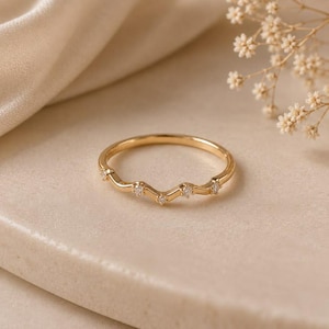 Puede incluir: Un anillo de oro con un diseño de banda delicada y ondulada. El anillo está engastado con cinco pequeñas piedras claras de corte redondo espaciadas uniformemente a lo largo de la sección superior curva, creando un brillo sutil en la superficie.