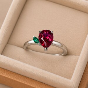May include: A silver-toned ring featuring a pear-shaped, deep red gemstone as the primary focal point, accented by a single, smaller marquise-cut green gemstone positioned to the side, both set on a polished, smooth, solid metal band.
