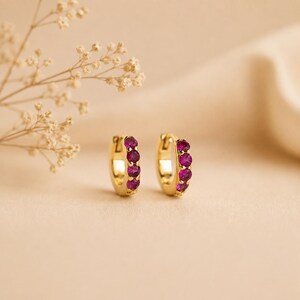 May include: A pair of gold hoop earrings, each featuring four small, round, vibrant magenta-pink gemstones set in a vertical row along the front curve of the hoop. The earrings have a polished metallic finish and a hinged closure mechanism.