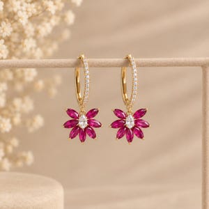 May include: A pair of gold hoop earrings featuring a row of small clear stones along the front. Each hoop suspends a flower-shaped charm composed of multiple marquise-cut fuchsia-coloured stones surrounding a single central clear oval-cut stone.