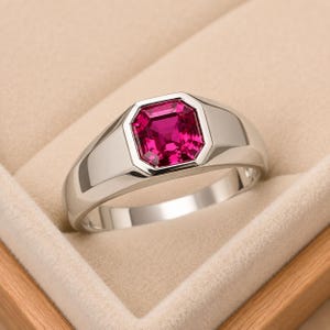 May include: A polished silver-tone ring featuring a vibrant, square-cut magenta gemstone set in a smooth, wide bezel. The band has a clean, solid construction with a high-shine metallic finish that reflects light across its curved surface.