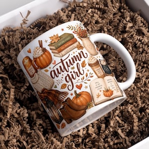 Op de afbeelding: Een witte keramische mok met een kleurrijke herfstillustratie en de tekst "autumn girl". Het ontwerp bevat gestapelde boeken, een pumpkin spice drankje, een kaars, een gebreide trui, bruine laarzen en een pompoen in warme herfstkleuren.