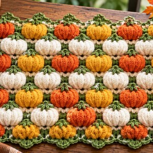 Op de afbeelding: Een rechthoekige gehaakte mat met een herhalend patroon van kleine, driedimensionale pompoenen in tinten oranje, geel en wit. De pompoenen zijn verbonden door groene gehaakte steeltjes en blaadjes, met een geschulpte groene rand rondom.