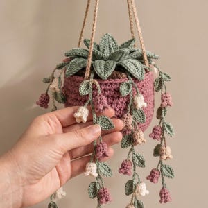May include: A crocheted hanging plant featuring a dusty rose pot, a central green succulent, and trailing vines with small bell-shaped flowers in shades of white, light pink, and dark pink, suspended by thin tan cords against a neutral background.