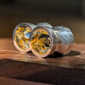 May include: Three silver-colored plugs with a polished, reflective finish. The plugs have a circular design with a swirling pattern of gold, brown, and blue hues. The plugs are displayed on a wooden surface.
