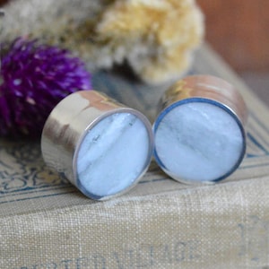 May include: Two round silver earrings with a white stone inlay. The earrings are sitting on a book with a floral design.