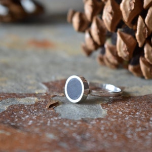 Puede incluir: Un anillo de plata con una piedra circular negra engastada en el centro. El anillo está sobre una superficie marrón texturizada.