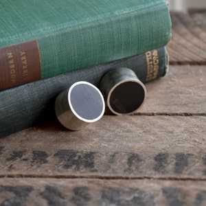 May include: Two black wood ear gauges with silver metal settings. The gauges are on a wooden surface.