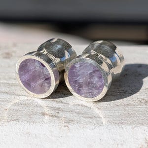May include: Two silver-colored plugs with a polished finish. Each plug features a circular, light purple amethyst stone set within the silver. The plugs are displayed on a textured, light-colored surface.