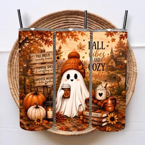 May include: A cylindrical tumbler with a straw featuring an autumn-themed design. The print displays a ghost wearing an orange beanie, pumpkins, a wooden sign with text, a house, and the words FALL VIBES AND COZY NIGHTS amidst fallen leaves.