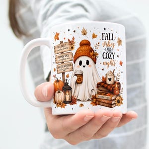 May include: A white ceramic mug featuring a ghost wearing an orange beanie, holding a coffee cup. The design includes a wooden signpost with text, pumpkins, a lantern, a stack of books, and the text FALL VIBES AND COZY NIGHTS in black font.