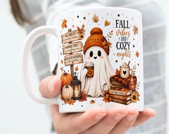 Könnte beinhalten: Ein weißer Keramikbecher mit einem Geist, der eine orangefarbene Mütze trägt und einen Kaffeebecher hält. Das Design zeigt ein Holzschild mit Text, Kürbisse, eine Laterne, einen Bücherstapel und den Schriftzug FALL VIBES AND COZY NIGHTS.