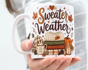May include: A white ceramic mug featuring the text Sweater Weather in brown script. The design includes illustrations of a stack of folded sweaters, a pumpkin, a small mug labeled cozy comfy happy, a candle labeled warm & cozy, and autumn leaves.