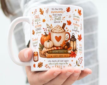 May include: A white ceramic mug featuring a colorful autumn-themed illustration of a pumpkin, a steaming cup of coffee with a heart, a lantern, and stacked books. The design includes text such as It's the little moments that make Fall special.