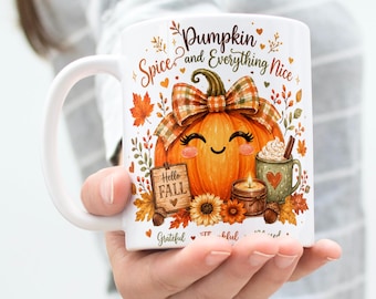 May include: A white ceramic mug featuring a colorful illustration of a smiling orange pumpkin with a plaid bow, surrounded by autumn leaves, a small sign reading Hello Fall, a candle, and a green mug with a heart, with text Pumpkin Spice and Everything Nice.