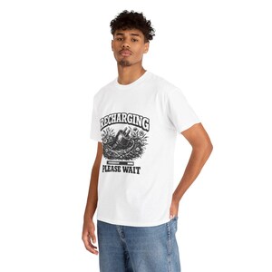 May include: A white short-sleeved t-shirt featuring a black graphic print on the chest. The design shows an electrical plug nestled in a bed of flowers with the text RECHARGING above and PLEASE WAIT below, along with a loading progress bar icon.