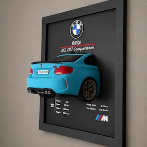 Könnte beinhalten: Ein gerahmtes Wanddisplay mit einem 3D-Modell eines hellblauen BMW M2 F87 Competition. Der schwarze Hintergrund enthält das BMW-Logo, technischen Fahrzeugdaten und ein Nummernschild mit dem Text YOUR TEXT in weißer Schrift.