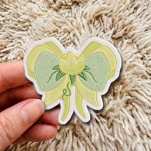 Può includere: Una toppa ricamata a forma di fiocco nei toni del verde chiaro e del giallo. Il centro del fiocco presenta un bocciolo di fiore stilizzato con foglie verde scuro e un piccolo viticcio che si estende verso il basso, bordato di filo bianco.