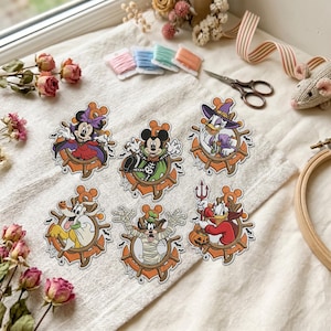 Op de afbeelding: Zes gestanste stickers met Disney-figuren in Halloween-kostuums, waaronder Minnie als vampier, Mickey als skelet, Daisy als heks, Pluto als geest, Goofy als mummie en Donald als duivel, allemaal omlijst door een scheepsroer.