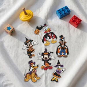 May include: A collection of six Disney character stickers featuring Mickey Mouse, Minnie Mouse, Donald Duck, Daisy Duck, Goofy, and Pluto dressed in Halloween costumes, including witch hats, capes, and pumpkin motifs, alongside a yellow spinning top.