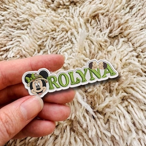 Op de afbeelding: Een geborduurde patch met de naam ROLYNA in groene letters met een witte omlijning. Aan de linkerkant staat een getekend muisfiguurtje met een safarihoed en een groene strik, en rechts een klein icoontje van een bruin dierenoor.