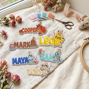 May include: A collection of six embroidered patches featuring cartoon characters and names. The patches include names Cooper, Mark, Leo, Maya, Sam, and Tony, paired with colorful illustrations of Vulpix, Diglett, Pikachu, Piplup, Squirtle, and Meowth.