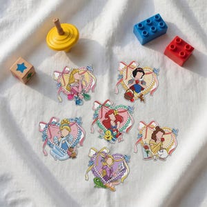 Può includere: Sei adesivi fustellati con personaggi illustrati incorniciati da bordi a forma di cuore con nastri e perle, accompagnati da una trottola gialla, un blocco da costruzione blu, un blocco rosso e un cubo di legno con una stella blu.