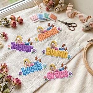 Puede incluir: Cinco parches bordados que muestran un personaje de dibujos animados con cabello rubio acostado sobre los nombres Abigail, Taylor, Rowan, Lucas y Grace. Cada parche incluye letras coloridas y pequeños detalles florales en naranja, morado, amarillo, azul y rosa.