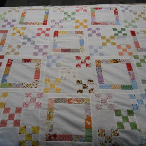 May include: A handmade quilt with a white background and a repeating pattern of squares and rectangles. The squares are bordered with colorful fabric patches in various patterns and colors, including red, blue, yellow, and green.