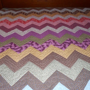 May include: A colorful quilt with a chevron pattern. The quilt features a variety of colors, including shades of brown, pink, purple, and blue. The quilt is likely made of cotton and is suitable for a bed or sofa.