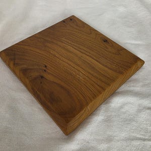 Può includere: Un tagliere quadrato in legno con una finitura naturale color marrone medio, caratterizzato da venature del legno visibili e piccoli nodi scuri su tutta la superficie. Il tagliere presenta bordi smussati e una superficie superiore piatta e liscia.