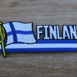 May include: An embroidered patch with the Finnish flag and the word "FINLAND" in white letters with a black outline. The flag is blue and white with a white cross on a blue background. The patch is on a wooden surface.