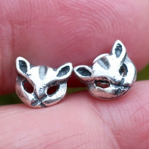 May include: Two small, silver-colored cat-shaped charms. Each charm features a detailed cat face with open eye and nose holes, and striped markings on the head. The charms are likely for jewelry making or crafting.