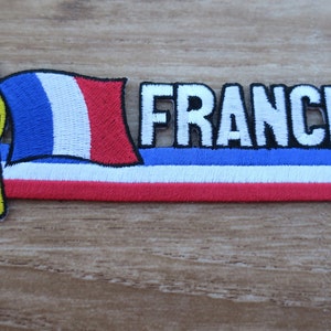 May include: Embroidered patch with the French flag and the word "FRANCE" in white letters with black outlines. The flag is on a pole with a yellow tassel.