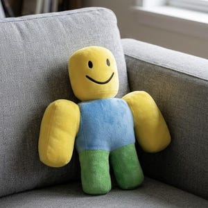 Puede incluir: Un peluche con forma de personaje de bloques con cabeza amarilla, una sonrisa y ojos negros simples, torso azul y piernas verdes. La figura tiene un diseño de cuerpo rectangular suave y redondeado con brazos gruesos y textura de tela.